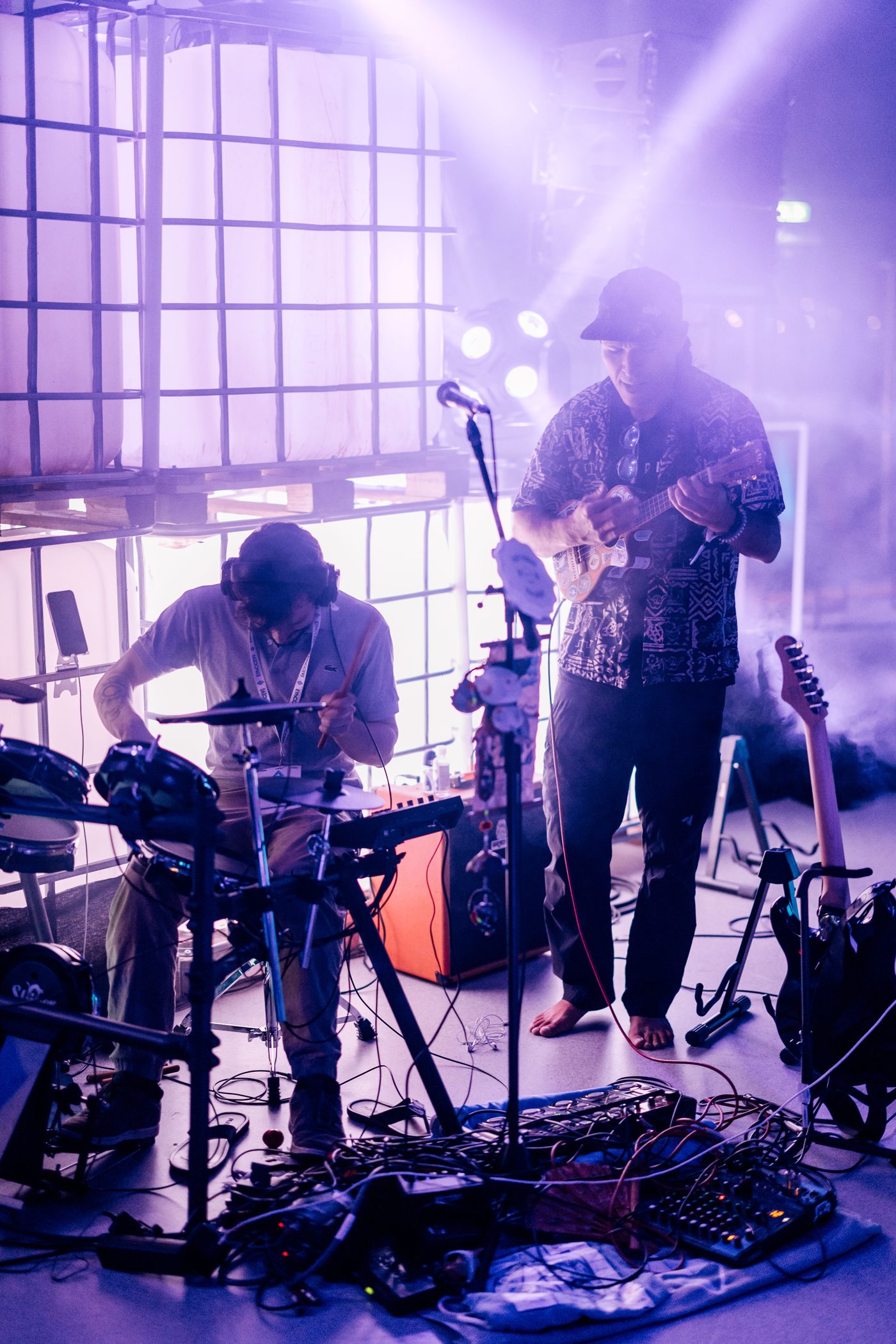 Shaka barefoot on ukulele, jamming with a drummer amid a sea of cables and synths